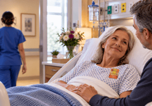 Older female patient in bed with an Elequil Aromatab on comfortably in bed with her husband and a nurse leaving the room