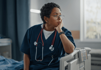african american nurse sitting at the end of a hospital bed with her head in her hand looking stressed