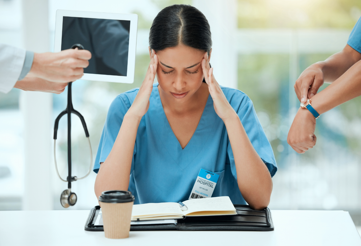 hospital tech sitting at a table holding her head with someone trying to hand her a stethoscope and someone else pointing at his watch