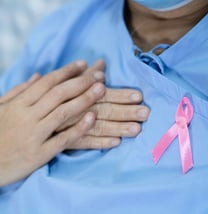 A hand holding a patient's hand over their chest next to a breast cancer ribbon pin.