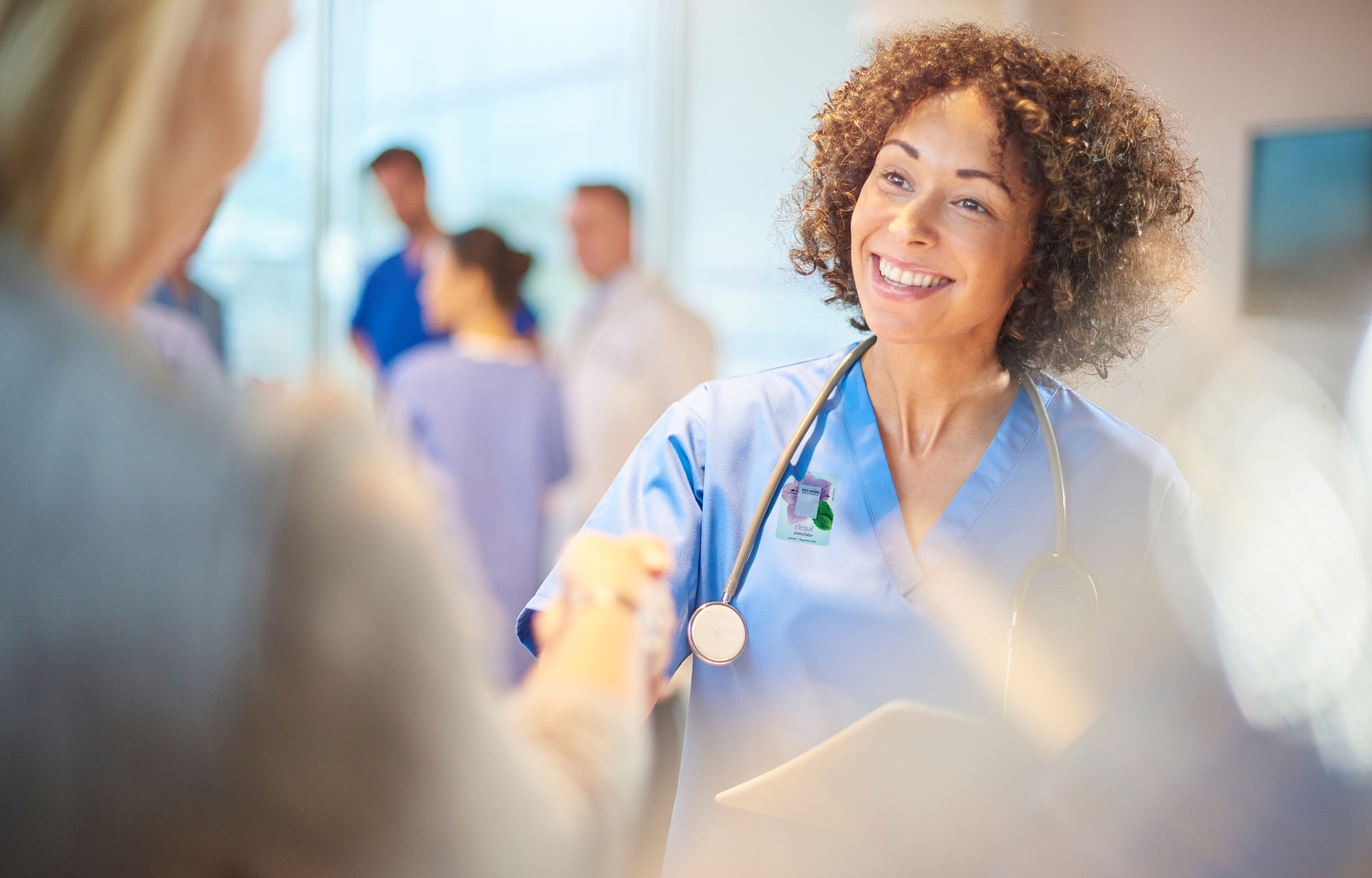 A nurse shaking someone's hand wearing an Elequil Aromatab on her scrub top.
