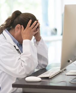 A doctor holding her head in her hands at a computer desk.
